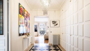 A hallway with a checkered floor, white walls, and a chandelier. There are artworks on the walls and a radiator on the right.