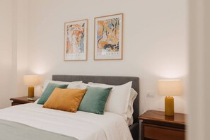Quarto de hotel com uma cama, duas mesas de cabeceira, duas luminárias e duas pinturas abstratas emolduradas na parede.