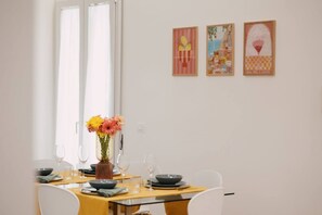 A dining area with a table set for four, a vase with flowers, and three framed artworks on the wall. - Elegant apartment in the heart of Syracuse, 3 bedrooms and 3 bathrooms, city center. (SIRACUSA)