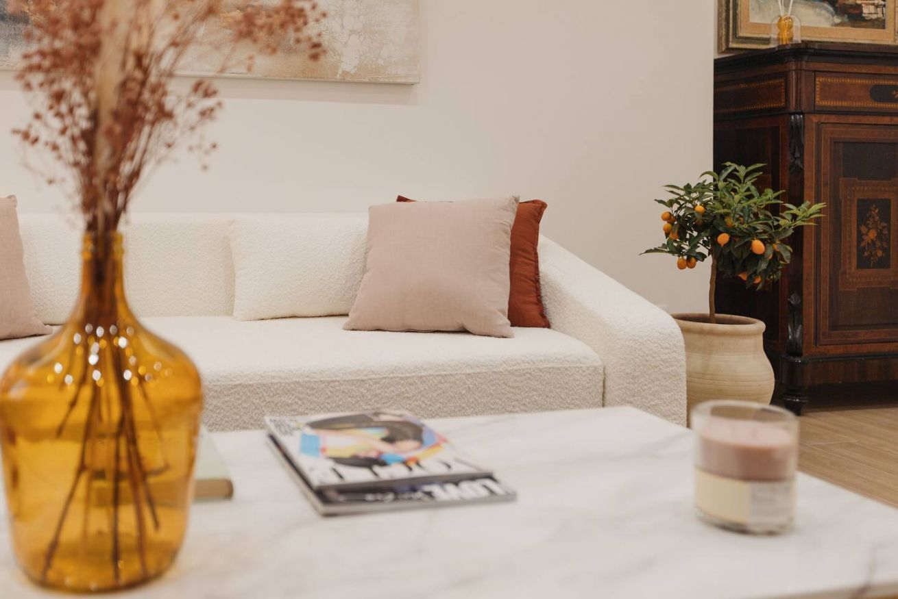 A white sofa with two pillows, a magazine, a candle, and a vase with dried flowers on a table.