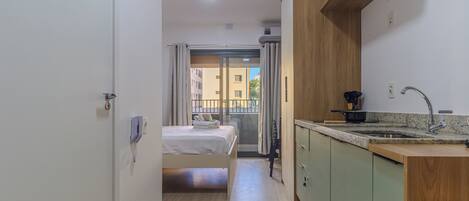 A compact hotel room with a bed, kitchenette, and a view through a sliding door to an outdoor balcony.