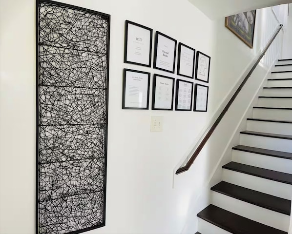 Eine Treppe mit schwarzen Stufen und einer weißen Wand. Sechs gerahmte Dokumente hängen an der Wand, darunter eines mit der Aufschrift 'wifi' und ein weiteres mit 'Nicht anfassen'.