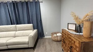 Un salon avec un canapé beige, des rideaux foncés, un meuble en bois orné d’objets décoratifs et un panier en osier.