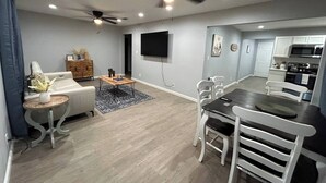 Un salon moderne comprenant un canapé, une table basse et un coin repas avec des chaises. On y trouve également un ventilateur de plafond, un éclairage encastré et un téléviseur à écran plat fixé au mur.