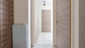 Un pasillo con un muro de mimbre, una puerta blanca y un piso de baldosas que conduce a una habitación con una puerta de madera y un aplique colgante.