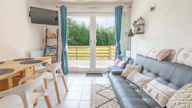 Una sala de estar moderna con un sofá gris, una mesa de comedor de madera con sillas, un televisor de pantalla plana y vistas a un jardín verde a través de una puerta de vidrio.