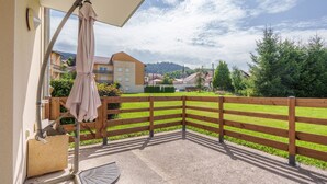 Un balcón con barandilla de madera, pared color beige y vista a un campo verde y edificios a lo lejos.