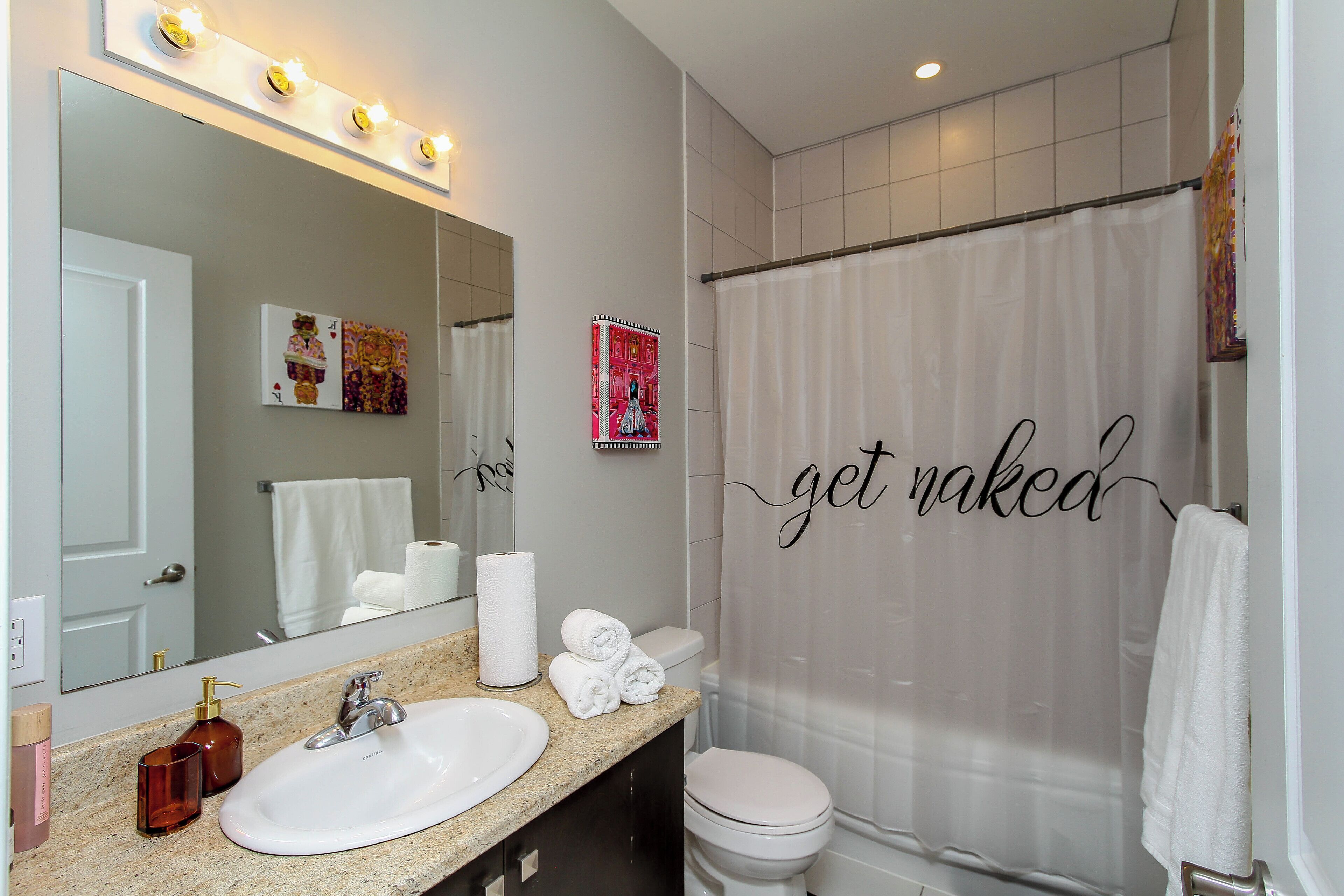 Een badkamer met een witte wasbak, toilet, douchegordijn met de tekst 'get naked' en een spiegel met verlichting.
