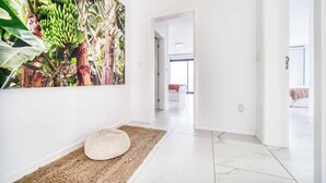 A hallway with a white wall, a large tropical-themed painting, a woven floor mat, and a glimpse of a bedroom with a bed and a window. - Casa das Flores - Three Bedroom Villa, Sleeps 6 (Calheta)