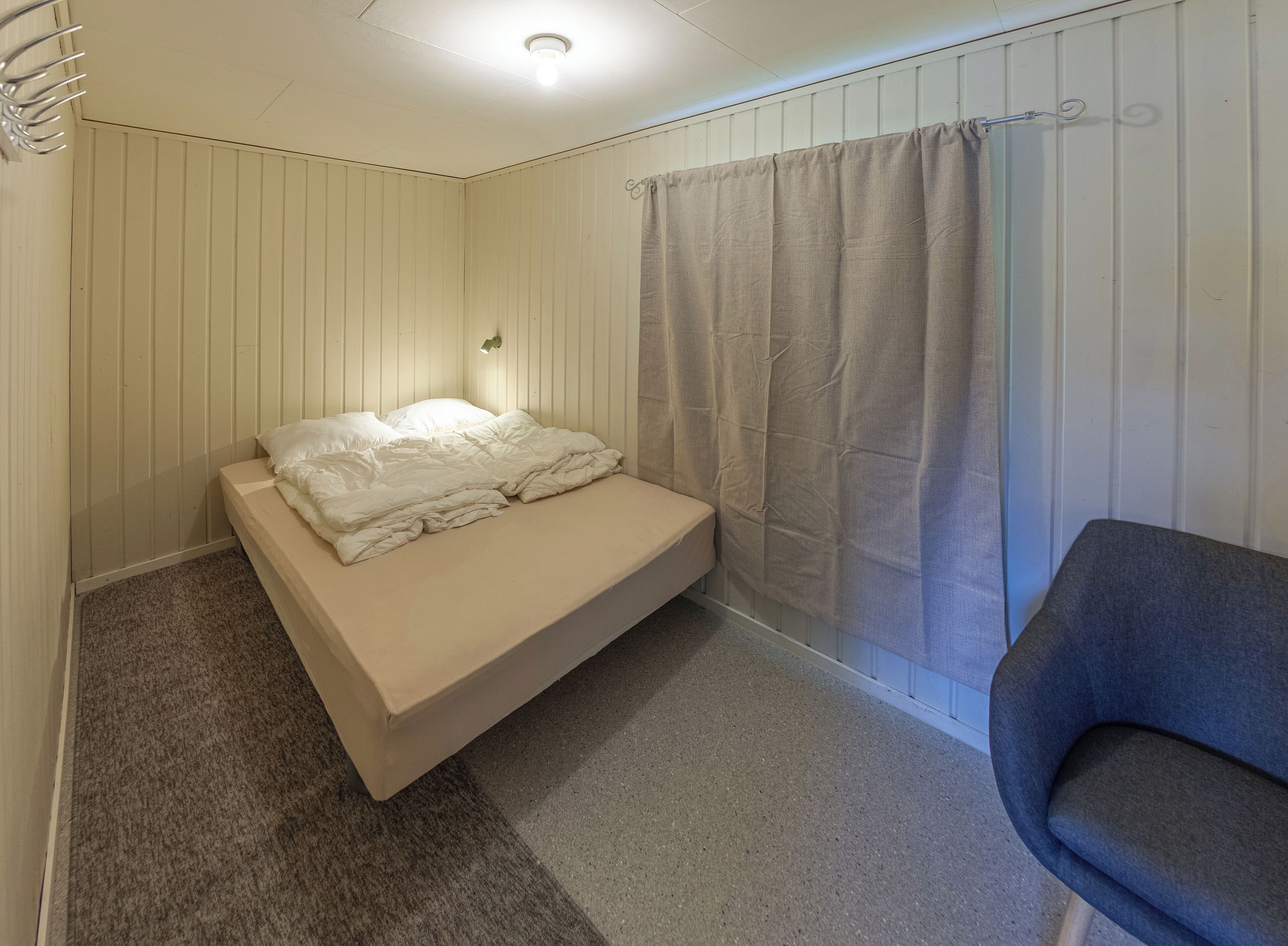 Ett litet rum med en enkel säng, en grå stol, ett fönster med gardin och en väggmonterad lampa.