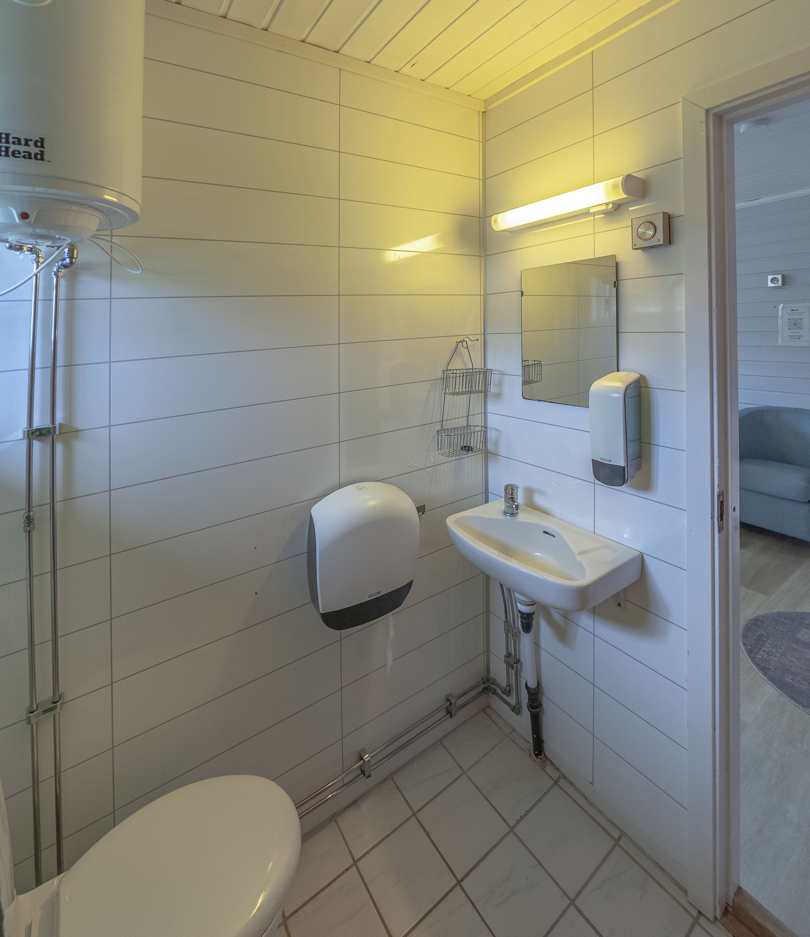 Une salle de bain avec des murs recouverts de carreaux blancs, des toilettes, un lavabo avec un miroir et un distributeur de savon.