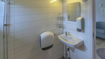 Une salle de bain avec des murs recouverts de carreaux blancs, des toilettes, un lavabo avec un miroir et un distributeur de savon.
