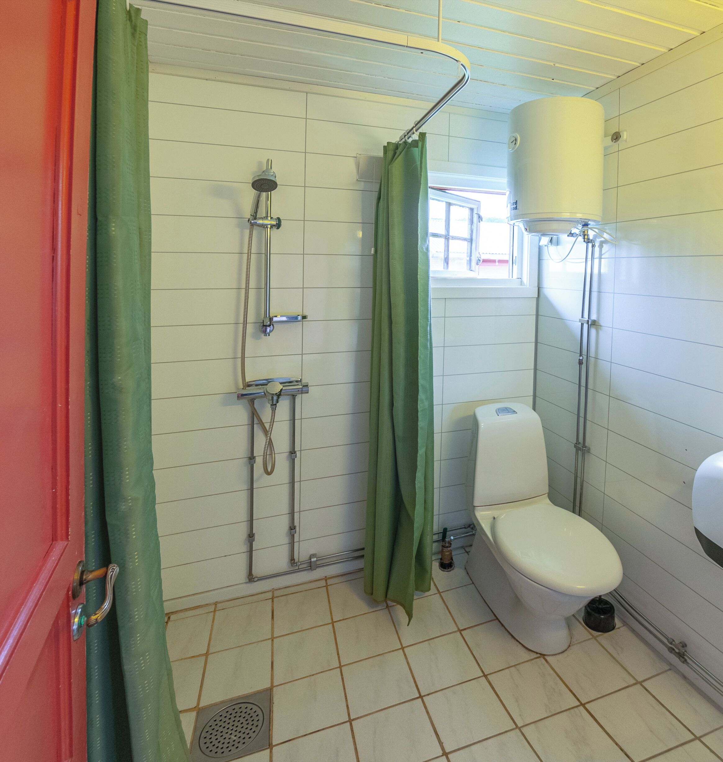 Une petite salle de bain carrelée, équipée d’une douche, de toilettes et d’une fenêtre.