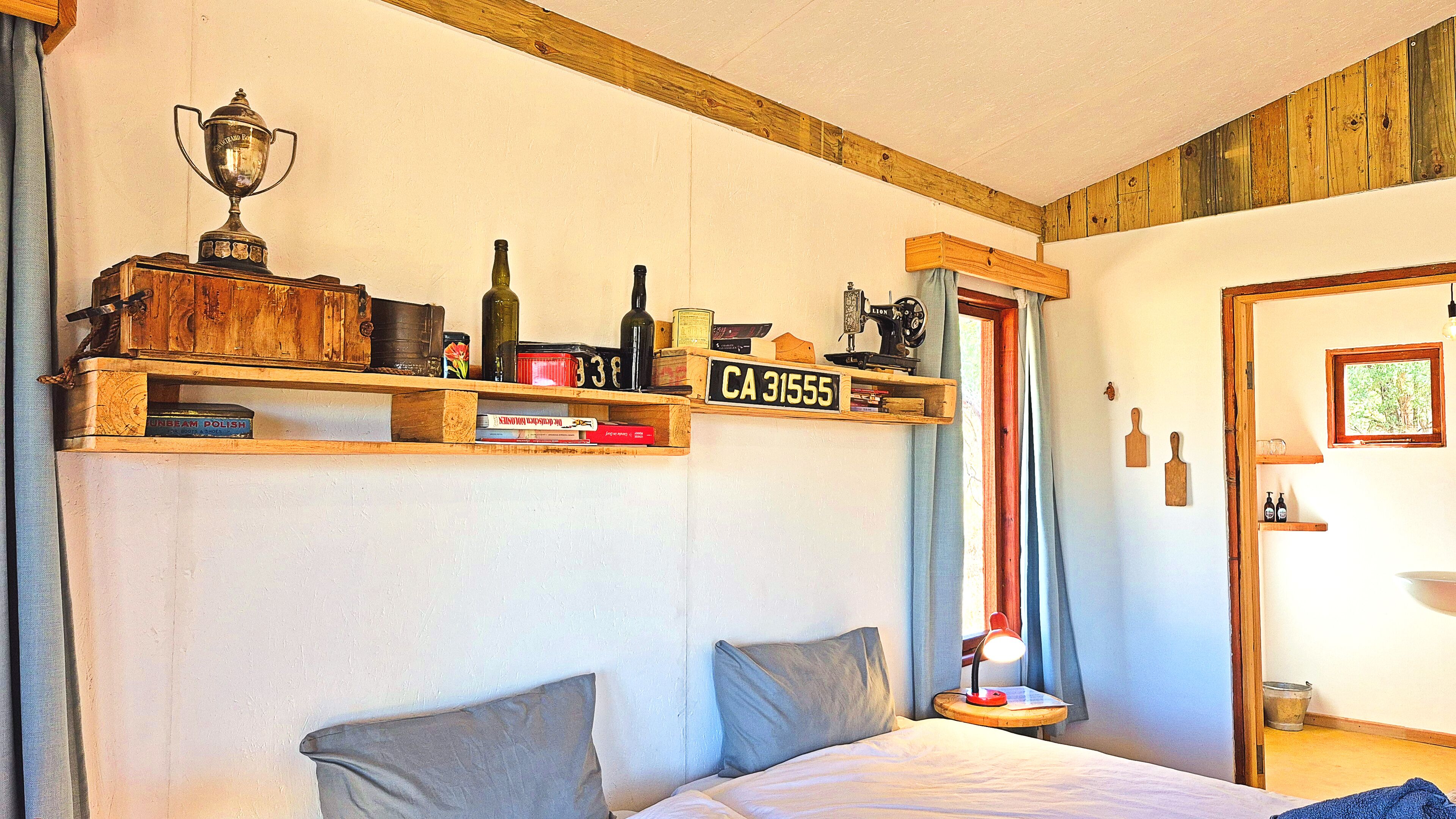 Een slaapkamer met een bed, kussens, een nachtkastje, een lamp, een trofee en houten planken met diverse spullen.