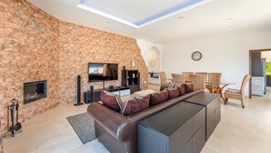 A modern living room with a stone wall, a large leather sofa, a flat-screen TV, and a dining area with wicker chairs.