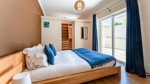 A bedroom with a wooden bed, a wardrobe, a small painting, and a view through a sliding door.