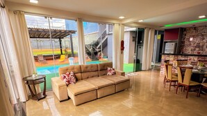 A spacious living area with a beige sectional sofa, a dining table with chairs, and a view of an outdoor swimming pool and stairs. - Clube Relax & Lazer (Foz do Iguaçu)