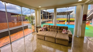 A spacious living area with a large window, a beige sofa, and a view of a swimming pool and outdoor seating area. - Clube Relax & Lazer (Foz do Iguaçu)