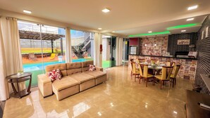 A modern living room with a beige sofa, dining area with chairs, a kitchen with a brick backsplash, and a view of a swimming pool and outdoor seating. - Clube Relax & Lazer (Foz do Iguaçu)