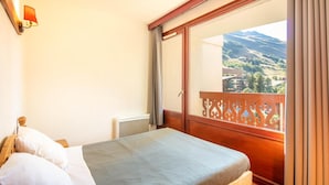 Una habitación de hotel con una cama, un ventanal con vistas a un paisaje montañoso y un balcón con barandilla de madera.