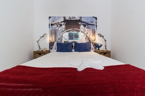 Un lit avec une couverture rouge, des oreillers blancs et une étoile de mer en guise de décoration. Il y a deux tables de chevet avec des lampes et un tableau représentant un bateau, accroché au mur.