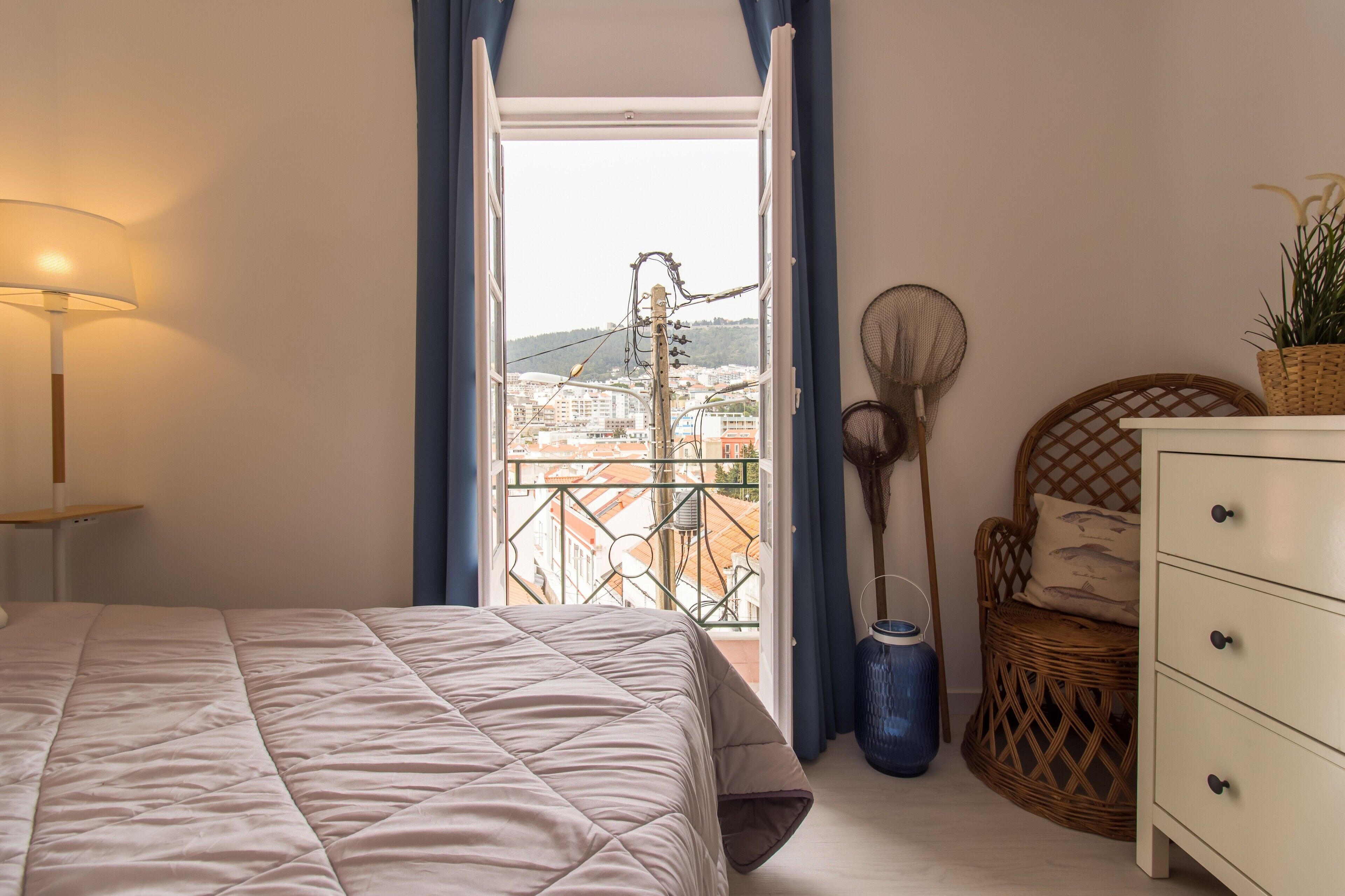 Un dormitorio con cama, cómoda, silla de mimbre, ventilador y vistas a edificios a través de una puerta de balcón.