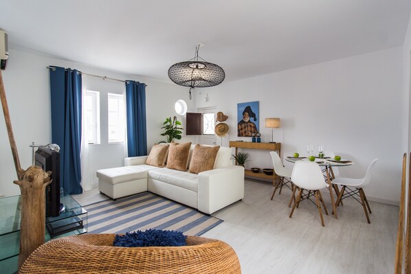 A modern living room with a white sofa, dining table, and chairs, a large window with blue curtains, and a hanging light fixture.