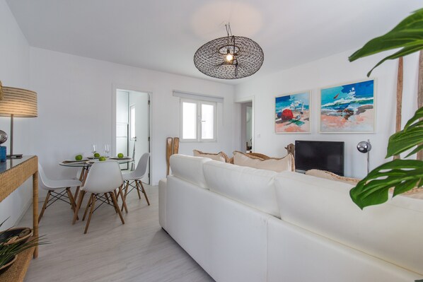 Un salon moderne avec un canapé blanc, une table à manger et des chaises, un téléviseur et des œuvres d’art accrochées au mur.