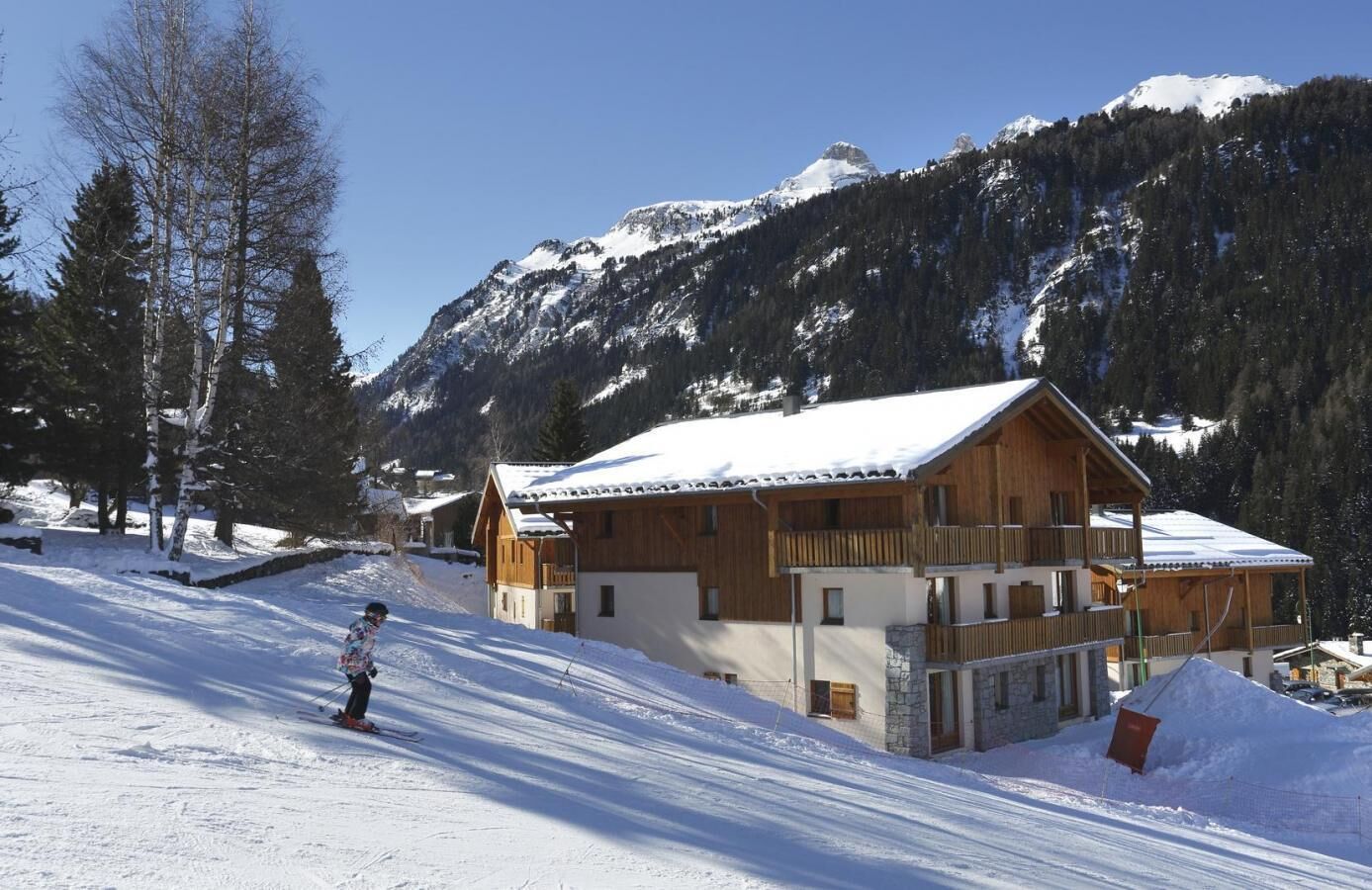 Een houten chalet met een sneeuwbedekte dak, tegen een achtergrond van bergen en bomen.