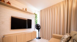 A modern living room with a flat-screen TV mounted on a white brick wall, a wooden cabinet below, a floor fan, and a beige sofa with patterned cushions. - Chaves do Atlântico (Cabedelo)