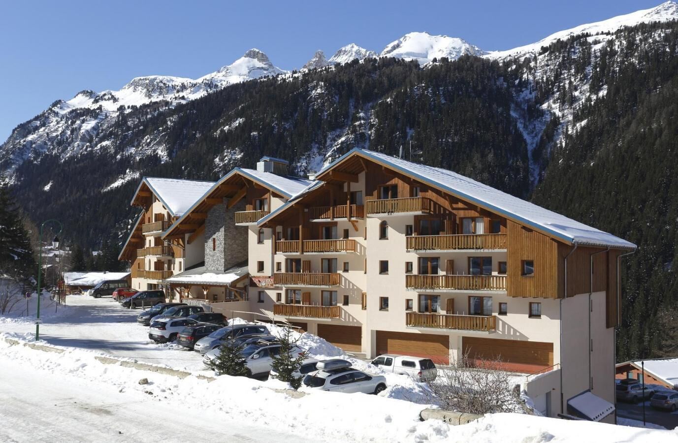 Une station de ski avec des bâtiments en bois, un parking et, en arrière-plan, des montagnes enneigées.