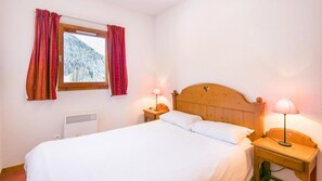 A neatly made bed with white linens, two bedside lamps, a window with red curtains, and a wooden headboard. - Wifi, 300m from resort center, swimming pool, sauna, tv, ski locker, 40m², Valfréjus (Valfréjus)