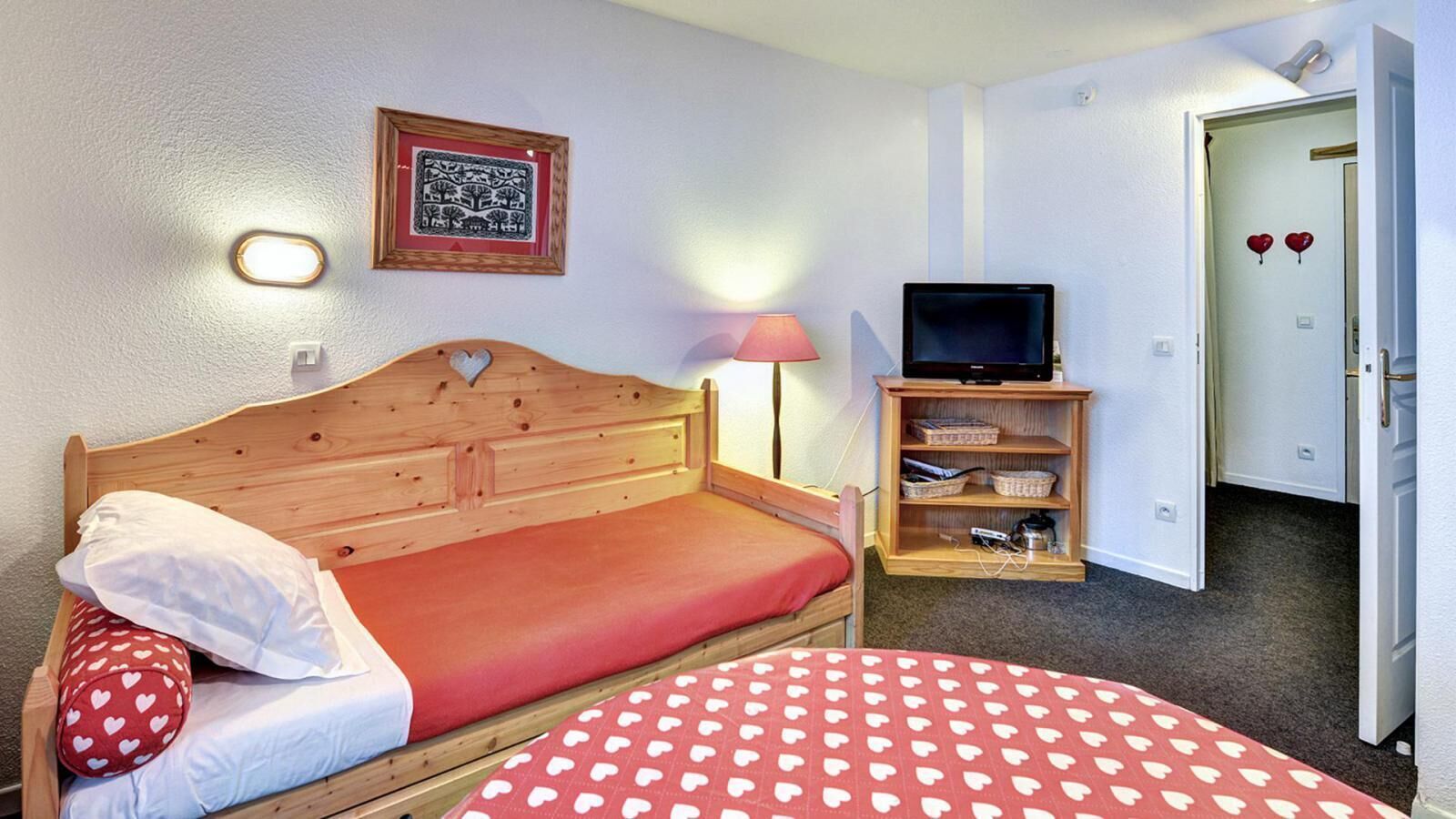 A bedroom with a wooden bed, a red heart-patterned bedspread, a bedside lamp, a television on a wooden stand, and a framed picture on the wall.