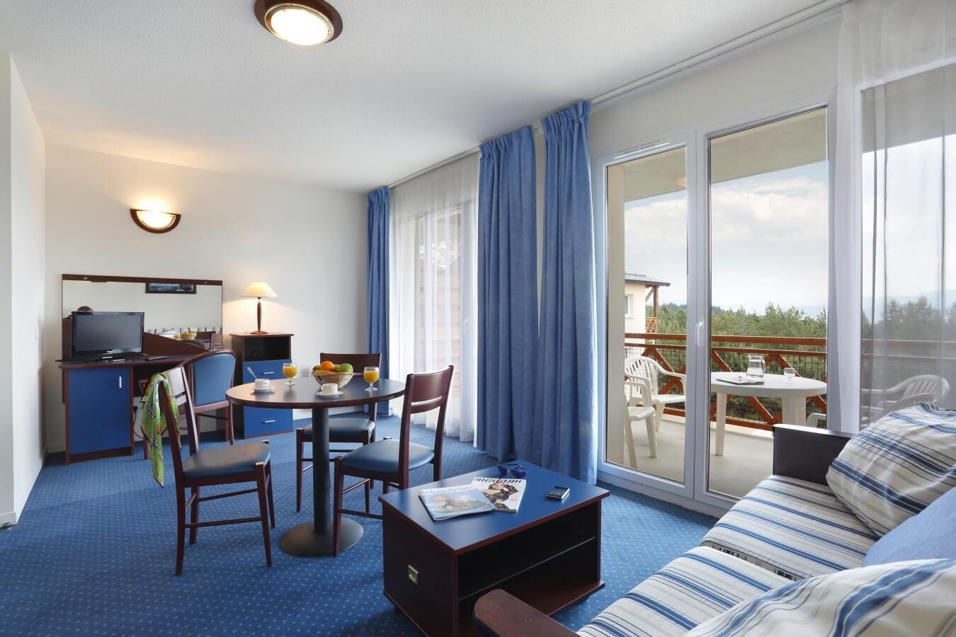 A hotel room with a dining area, a sofa, a TV, and a balcony with a view.