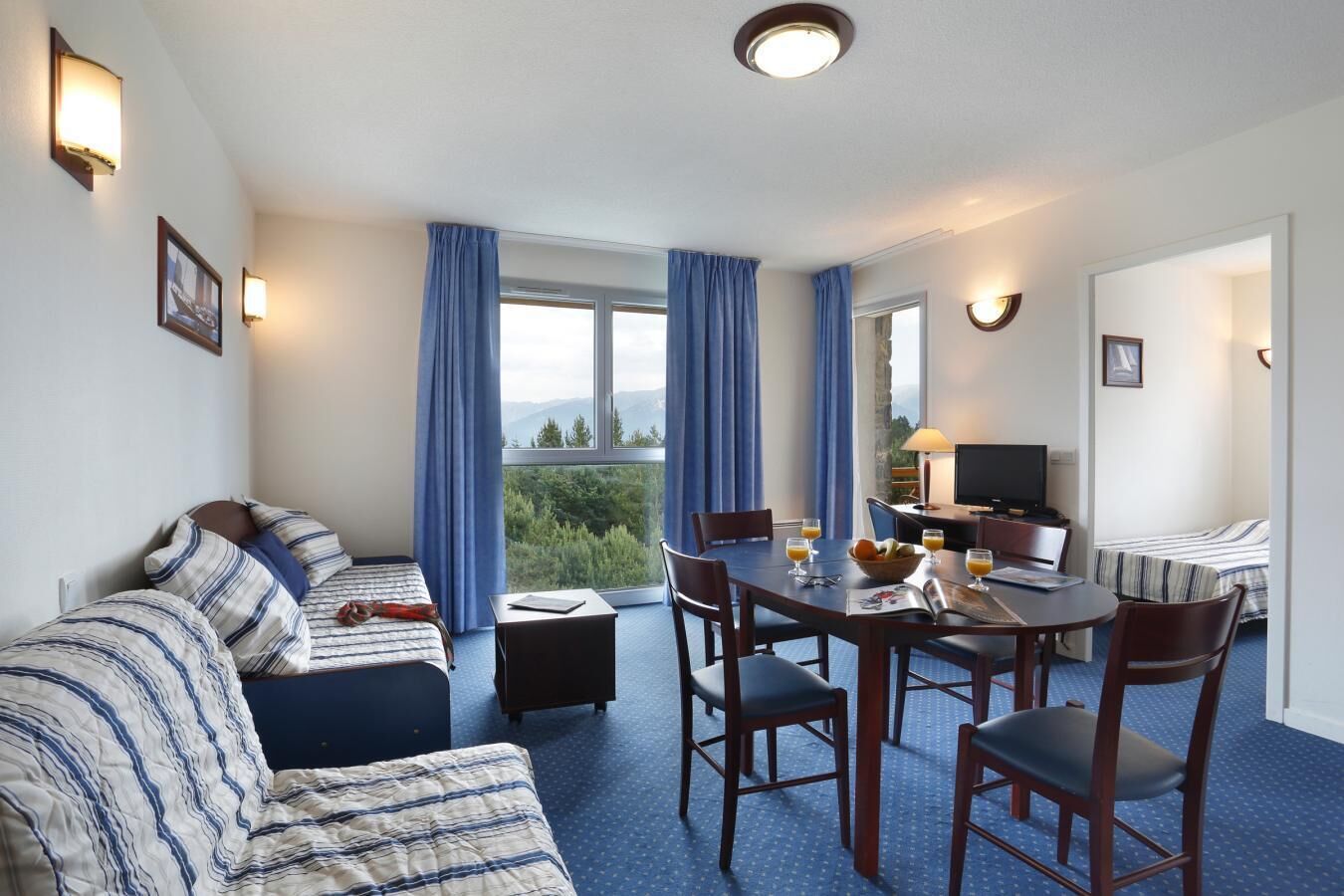 A hotel room with a bed, a sofa, a dining table with chairs, a TV, and a window with curtains.