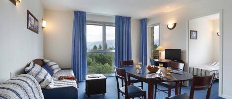 A hotel room with a bed, a sofa, a dining table with chairs, a TV, and a window with curtains.