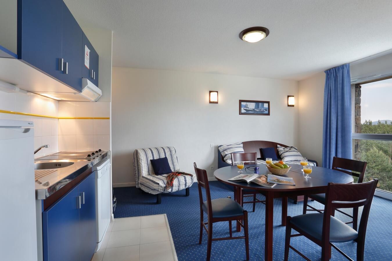 A modern kitchen with a built-in stove and sink, a dining area with a round table and chairs, and a balcony with a view.