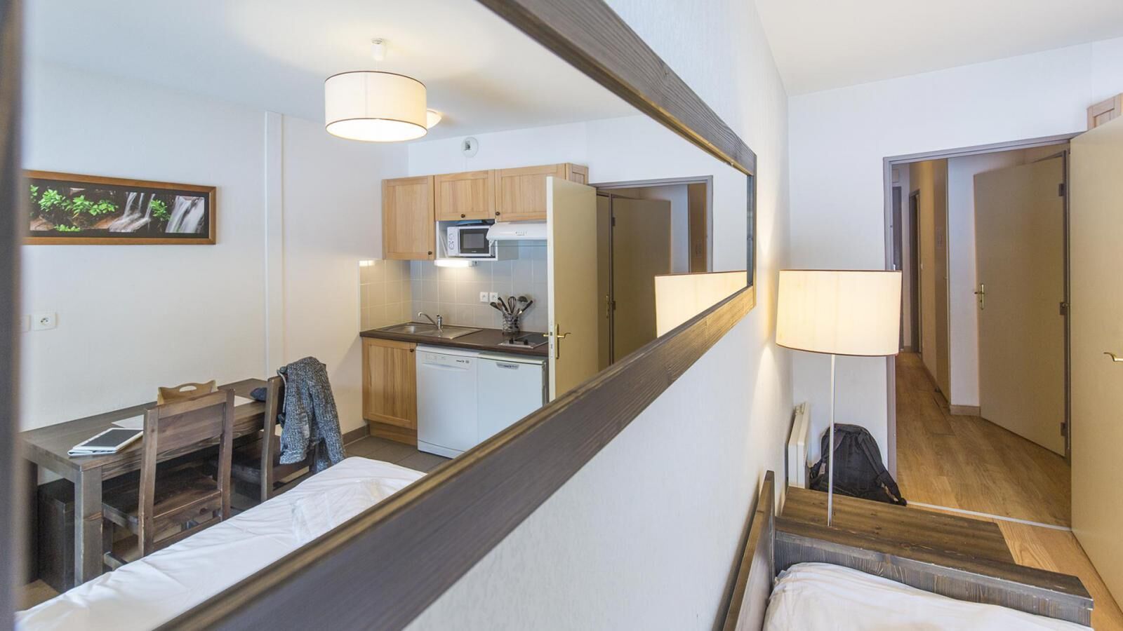 A compact hotel room with a bed, a desk, a chair, a small kitchenette, and a mirror.