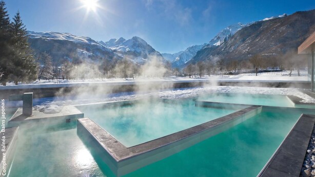 Une piscine d’eau cristalline, d’où s’échappe de la vapeur, entourée de montagnes enneigées et surplombée par un ciel d égagé.