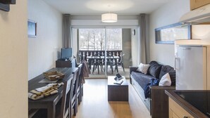 Un espace de vie compact comprenant un canapé, une table à manger et des chaises, une petite kitchenette et une vue sur l’extérieur grâce à une porte vitrée.