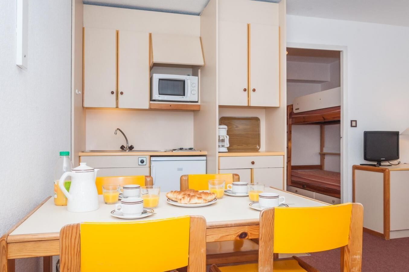 Een kleine kitchenette met een magnetron, koffiezetapparaat en een kleine koelkast. Er is een eettafel met gele stoelen, en een stapelbed met een televisie in de hoek.