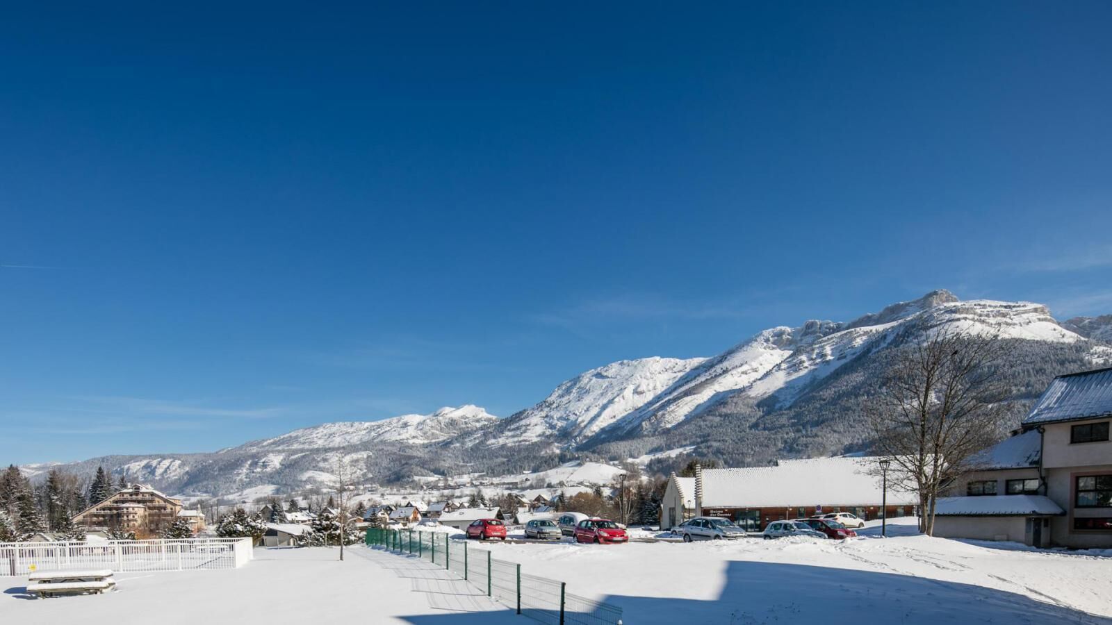A snowy mountain town with a clear blue sky, a parking lot with several cars, and a prominent mountain in the background.
