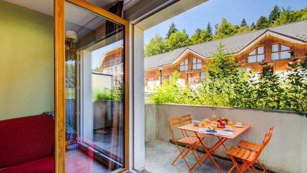 A balcony with a table and chairs, a red sofa, and a view of wooden buildings and greenery. - 300m from ski slopes, swimming pool, parking, tv, ski locker, 40m², La Féclaz (Les Déserts)