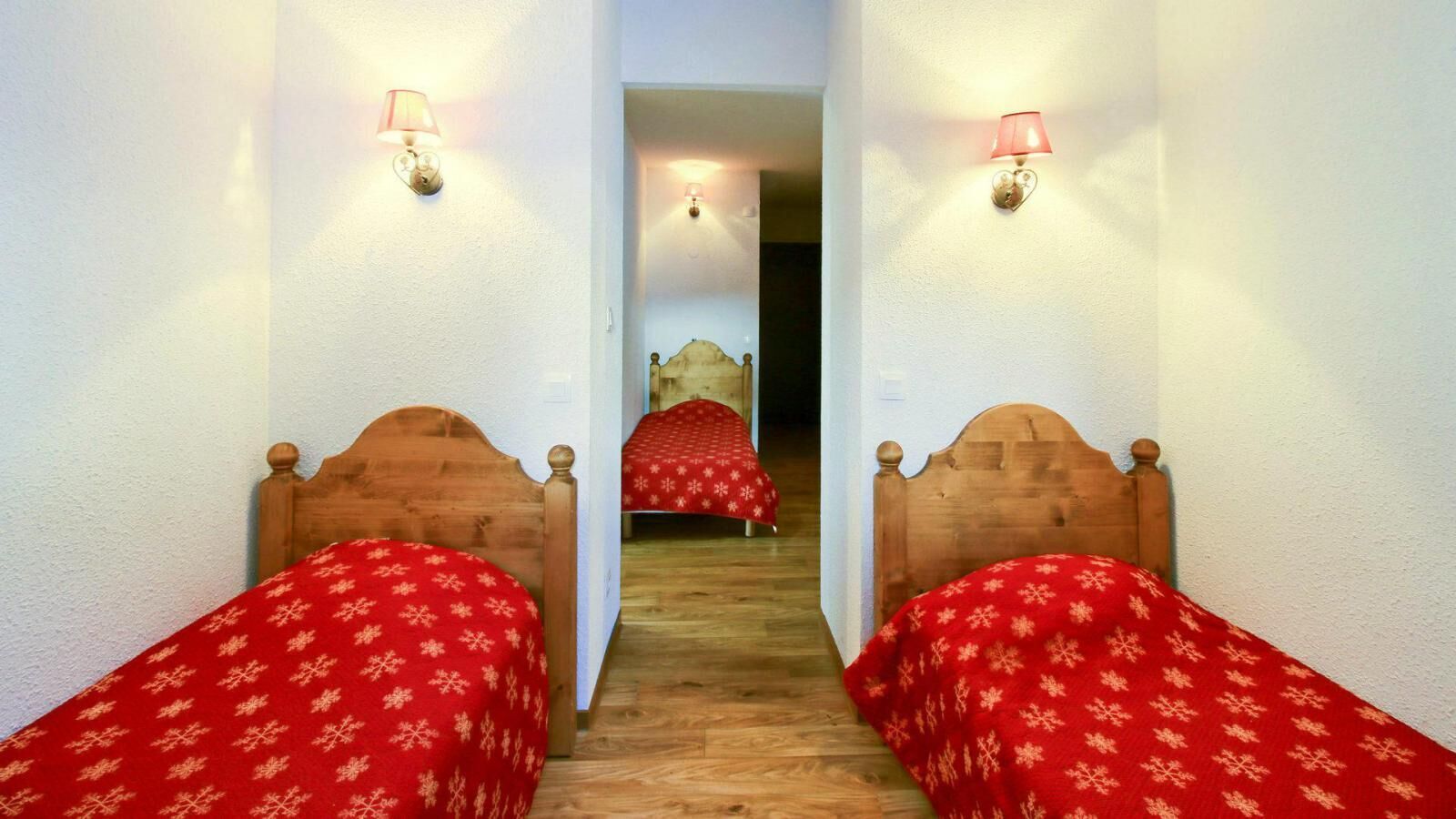 A hallway with two wooden beds, each with a red bedspread and a wall-mounted lamp.