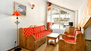 Ein gemütliches Zimmer mit einer Holzsofa und Sesseln, einem kleinen Tisch, einem Fernseher und einem Fenster mit Blick auf Gebäude und Bäume.