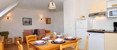 Un rincón comedor acogedor con una mesa de madera dispuesta para cuatro personas, rodeada de sillas. Se ve un refrigerador y un microondas blancos en la zona de la cocina a la derecha.