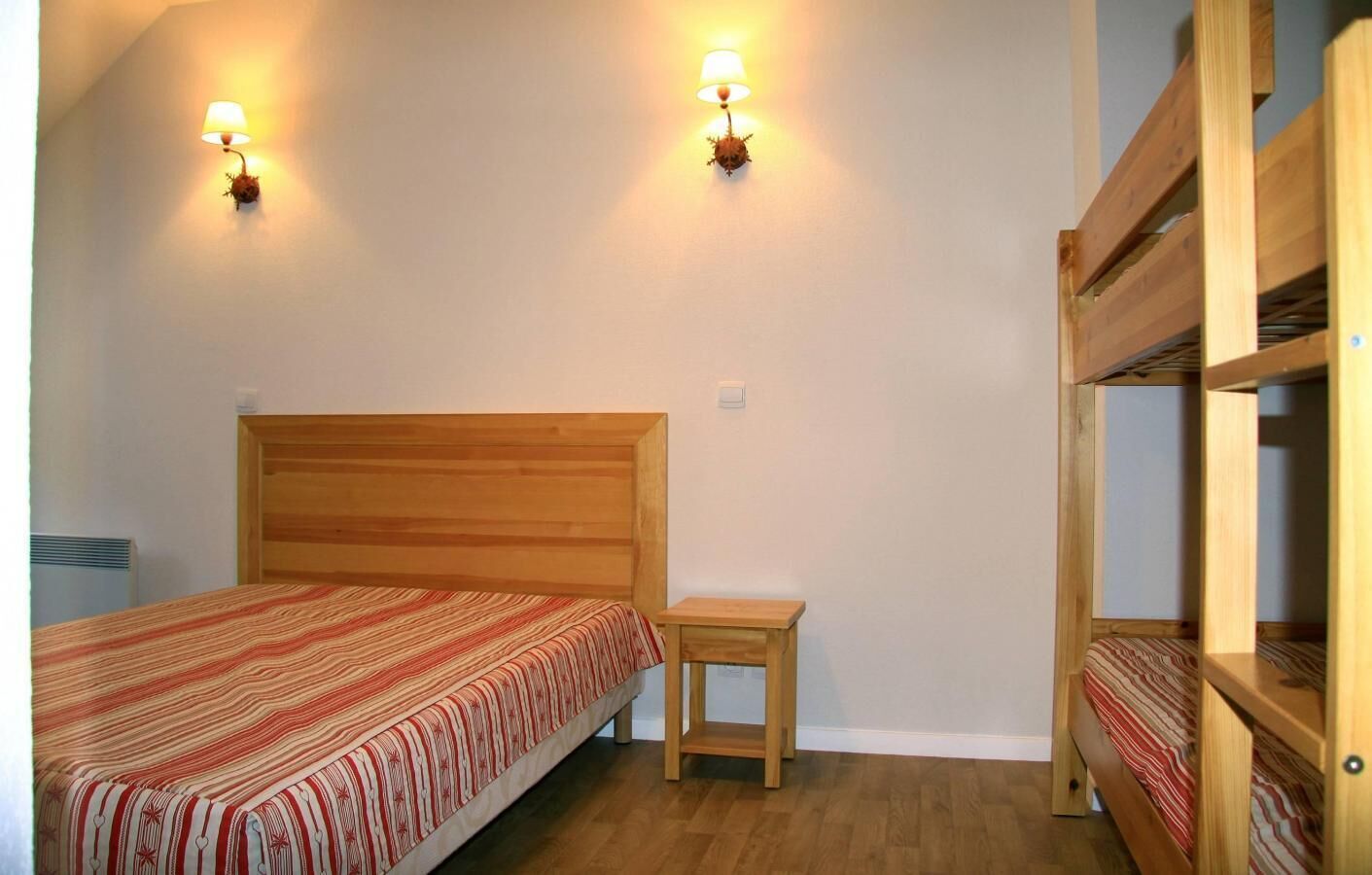 A bedroom with a wooden bed, a small bedside table, and a bunk bed. There are two wall-mounted lamps providing light.