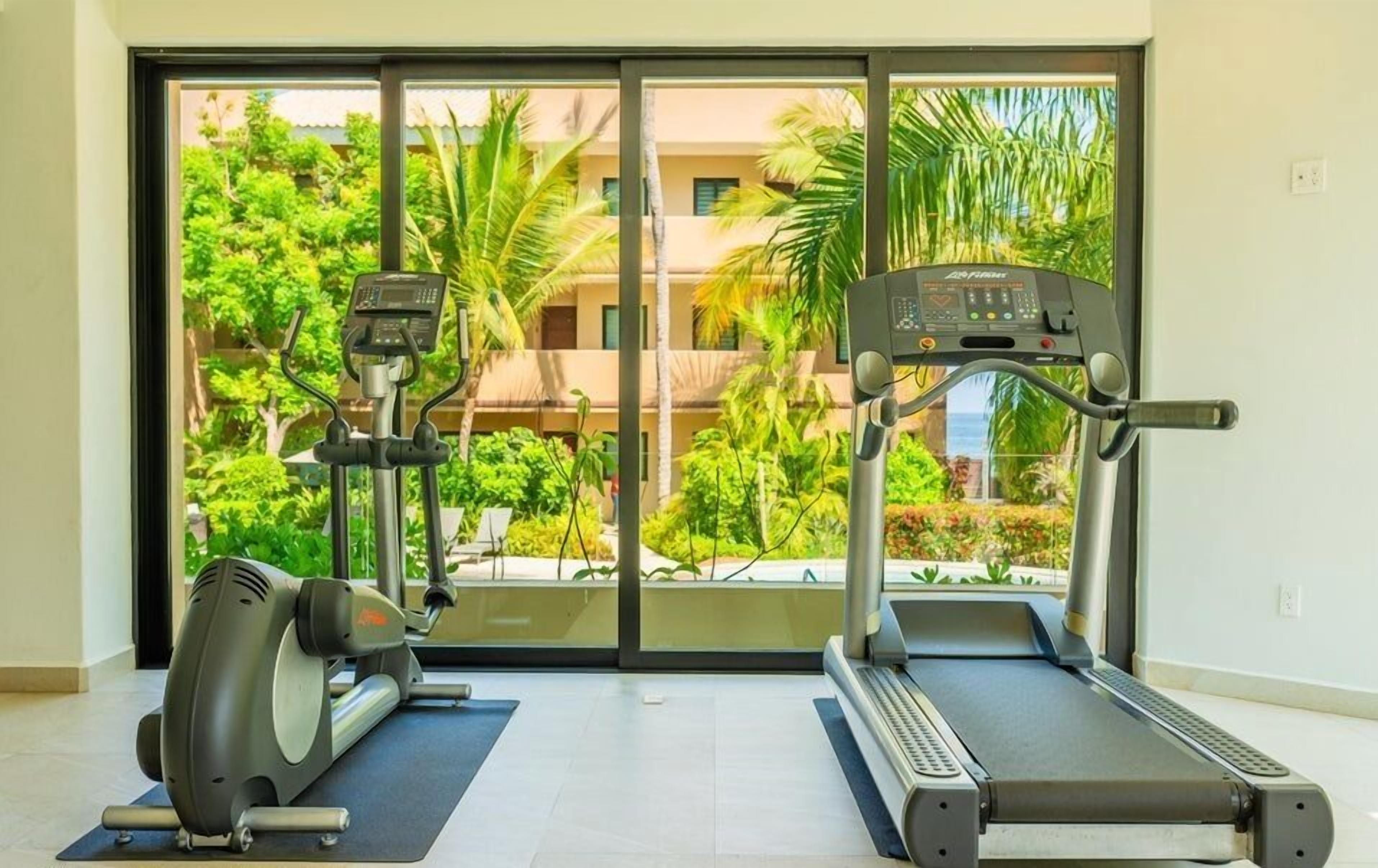 A gym with two treadmills and an elliptical machine, large windows with a view of greenery and buildings.