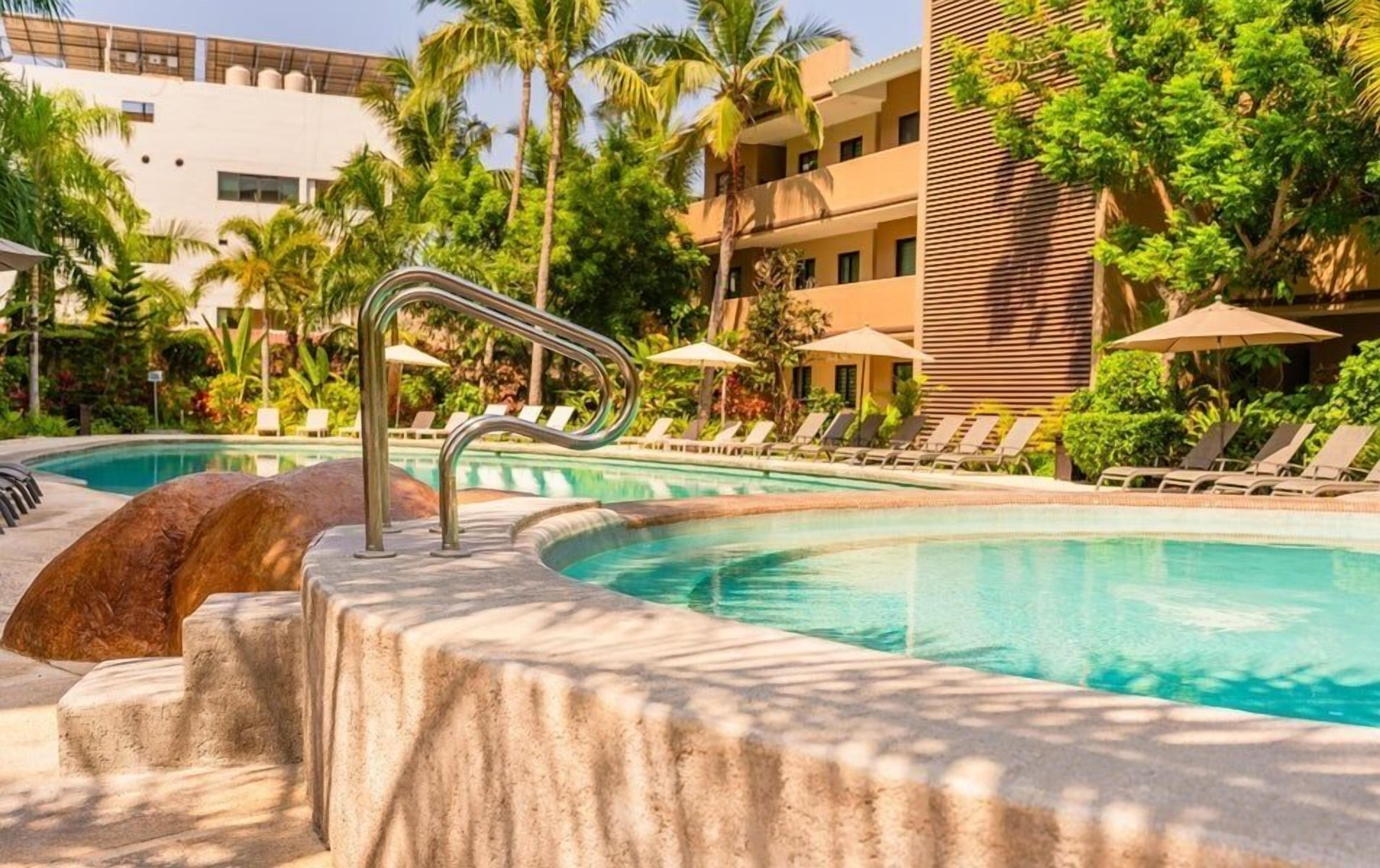A pool area with lounge chairs and umbrellas, surrounded by palm trees and a building with a modern design.