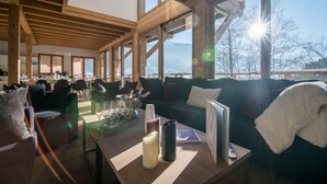 Tilava huone, jossa on korkea katto ja suuret ikkunat, sohva, lasien ja kynttilän koristama sohvapöytä sekä näkymä ulos.